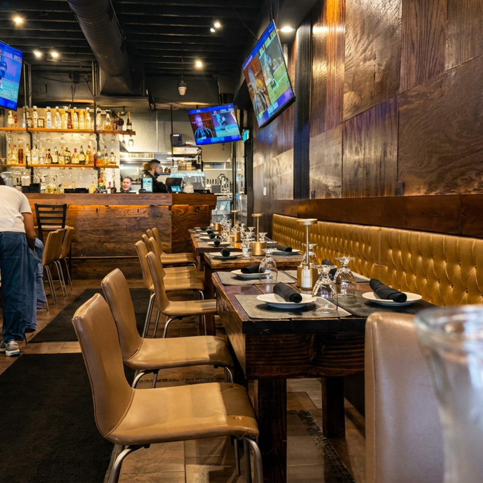 Warmly lit restaurant dining room with leather booths, set tables, wooden bar, liquor shelves, televisions, and a staff member.